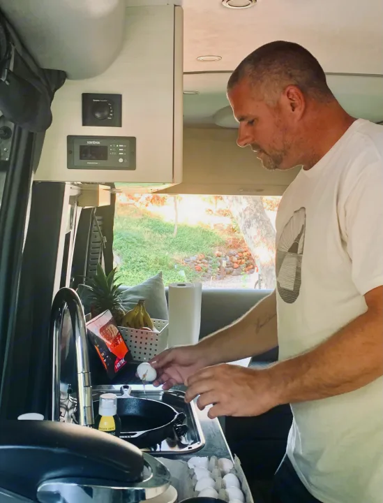Man cracks egg into pan in a small RV kitchen with controls above. Perfect for an rv rental adventure.. Pleasure-Way Pleasure-Way Class B 2020