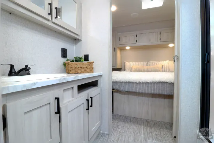 This RV rental features a modern light wood bathroom vanity, sink, and a bright bedroom visible through the doorway.. East to West Silver Lake 2023