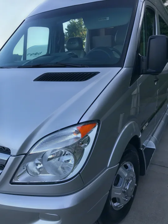 This silver camper van for rv rental shows the front passenger side with its headlight, hood, and shiny wheel.. Coachmen Galleria 2020