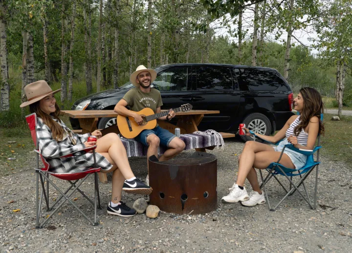 Friends enjoy guitar music, laughter, and drinks at a wooded campsite, near a black vehicle, suggesting a fun RV rental getaway.. Glacier Campers Cosy CrewVan 2015