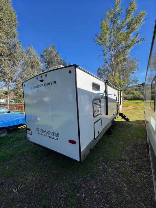 RV rental: A white travel trailer is parked outdoors on grassy ground under a clear blue sky, surrounded by trees.. Forest River Cherokee Alpha Wolf 2025