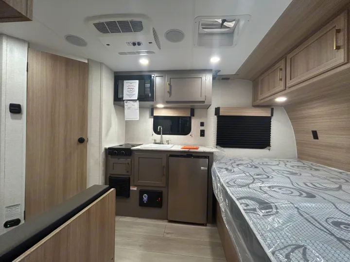 RV rental interior: compact kitchen with microwave & fridge, bed with plastic covering, and dinette. Modern wood-look finishes.. K-Z 130RD 2026