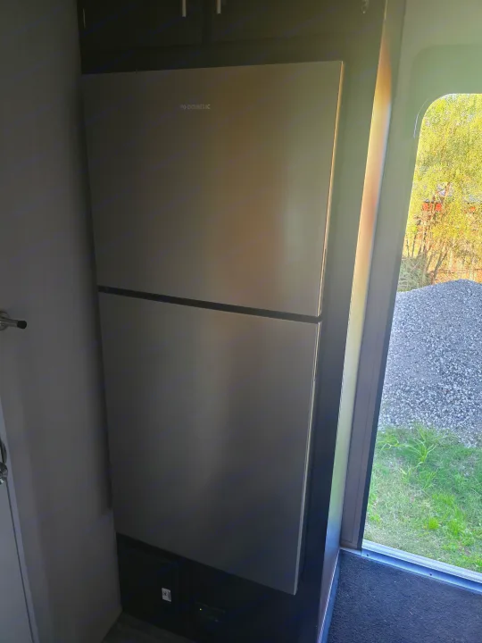 Stainless steel Dometic refrigerator in an rv rental, beside an open door with a sunny outdoor view of grass.. Dune Sport Dynamite 2023