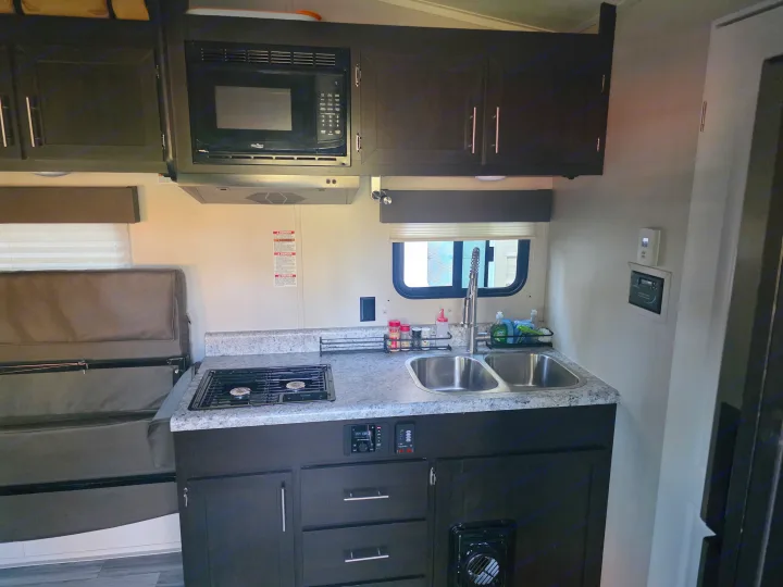 Compact rv rental kitchen with dark cabinets, microwave, Dometic stove, double sink, and a comfortable dinette seating area.. Dune Sport Dynamite 2023