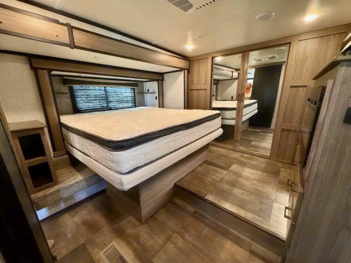 Modern RV rental bedroom with a spacious, raised bed, mirrored closet, warm wood cabinetry, and a step-up floor.. Crossroads RV Cameo 2021