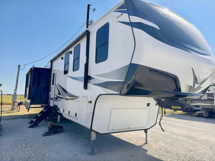 Large white fifth-wheel RV rental parked on gravel, with a dog resting by its steps under a clear sky.. Crossroads RV Cameo 2021