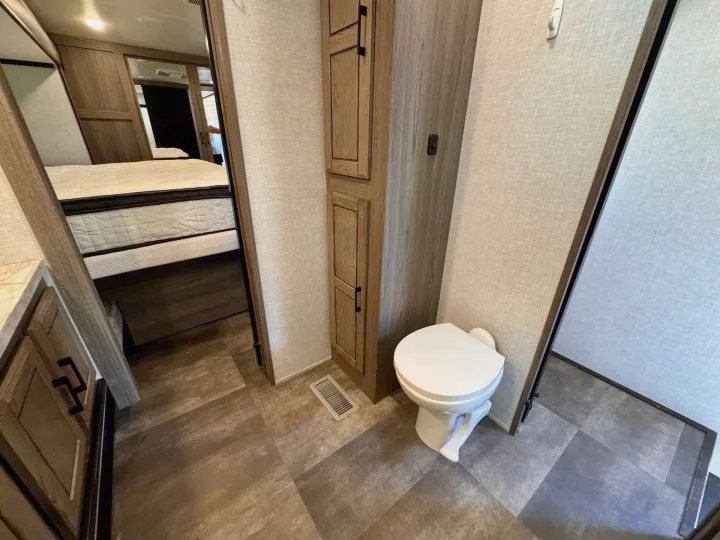 RV rental interior: A compact bathroom with a white toilet, wood cabinet, and a view into the adjacent bedroom.. Crossroads RV Cameo 2021