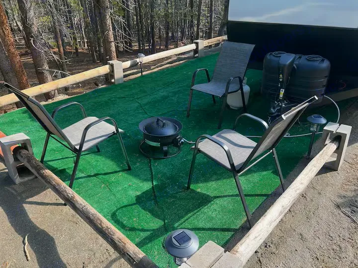 Cozy outdoor rv rental setup with chairs around a fire pit on green turf, nestled in a forest with an RV nearby.. Ember E 2024
