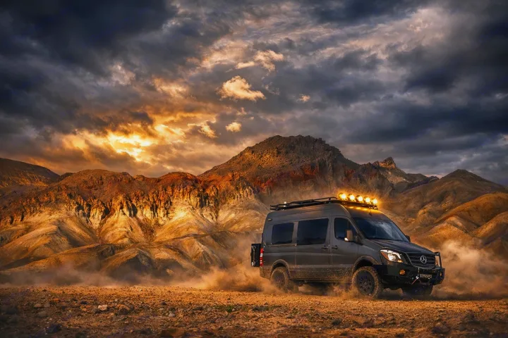 This rugged off-road Mercedes Sprinter van, perfect for an RV rental, kicks up dust in a dramatic desert landscape with epic mountains and a golden sky.. Sportsmobile 2500 2017