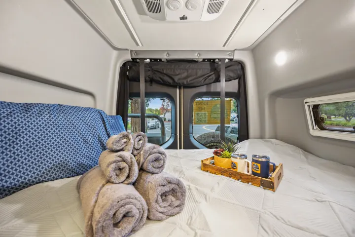 Cozy rv rental bed with blue pillows, rolled towels, and camping mugs on a tray. Perfect for your next adventure!. Winnebago Revel 2023