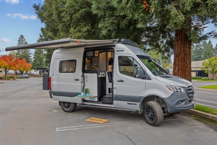 Silver Winnebago camper van rv rental with open side, extended awning, and outdoor dining setup in a parking lot.. Winnebago Revel 2023
