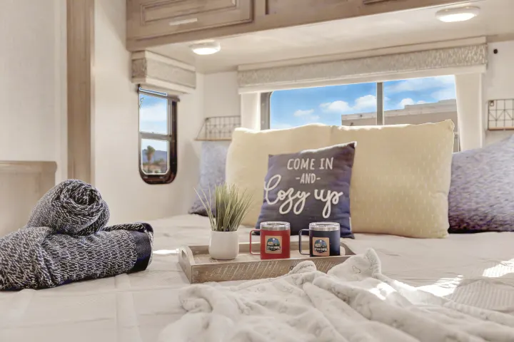 Cozy RV rental bedroom with 'Come in and Cozy up' pillow, blankets, plant, and 'Camping' mugs on a tray by sunny windows.. Forest River Georgetown 2019