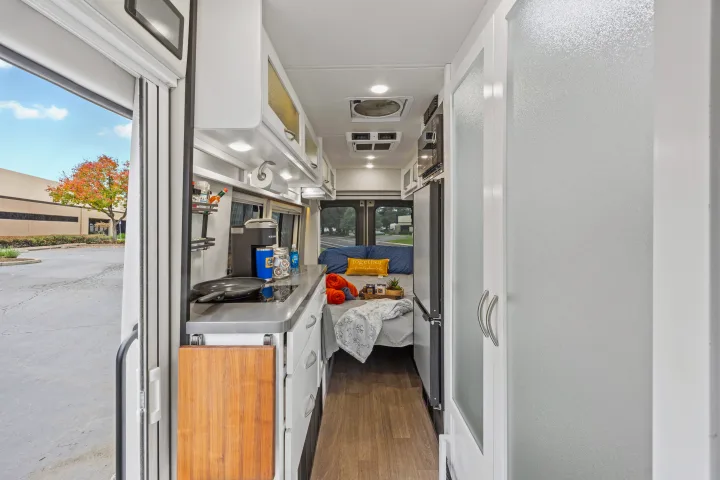 Bright rv rental interior with a kitchen, cozy bed, and frosted doors leading to a bathroom or storage. Autumn view outside.. Coachmen Beyond 2021