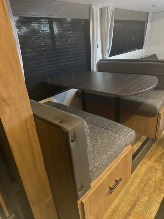 Experience comfort in this RV rental dinette, showcasing a dark wood table, grey booth seating with storage, and dark pleated window blinds.. Forest River Campsite Reserve 26CJ 2025