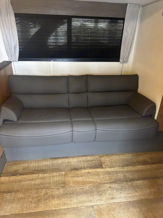 A comfortable dark grey sofa inside an RV rental, featuring a window with dark blinds and light wood-look flooring.. Forest River Campsite Reserve 26CJ 2025