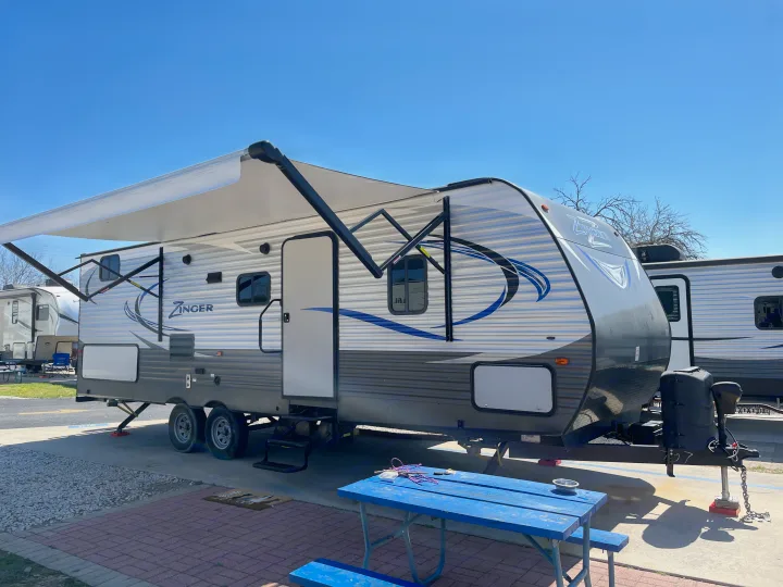 A white and gray travel trailer, an ideal rv rental, with an extended awning and a blue picnic table under clear skies.. Crossroads RV Zinger 2017
