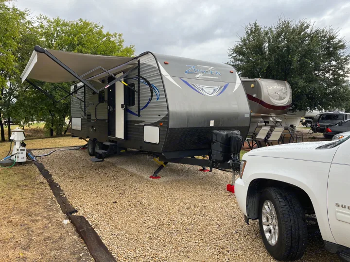 Zinger travel trailer RV rental parked at a campsite, with an extended awning & SUV. Utilities connected.. Crossroads RV Zinger 2017