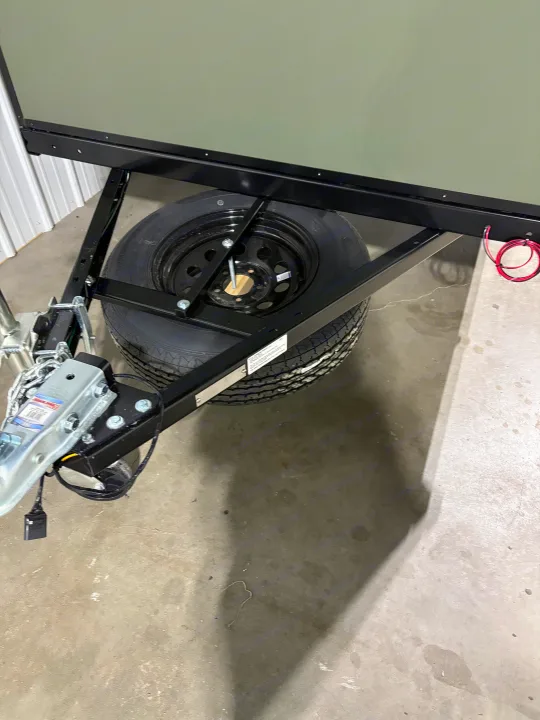 Front section of an RV rental trailer, showcasing the hitch, black chassis, and spare tire mounted underneath on a concrete floor.. Outbound Trailer Outbound Extreme 2026