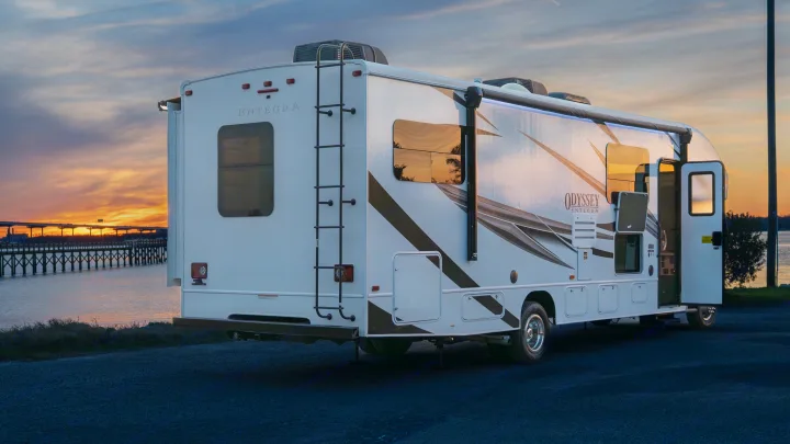 White Entegra Odyssey rv rental parked by a bay at sunset, door open, ladder on back. Perfect for travel.. Entegra Coach Odyssey 2023