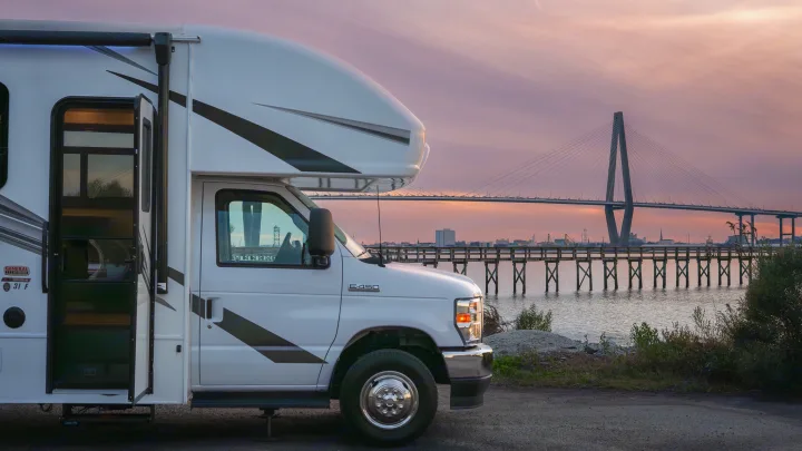 An RV rental with an open door is parked by a scenic waterfront with a large cable-stayed bridge and wooden pier.. Entegra Coach Odyssey 2023