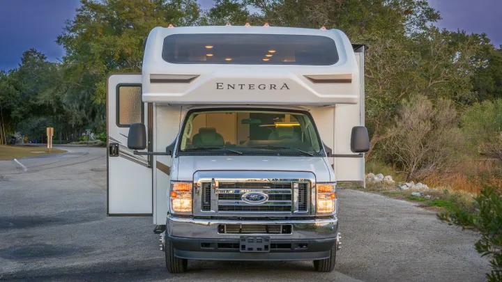 White Entegra Class C RV rental, headlights on & door open, parked on a winding road among trees.. Entegra Coach Odyssey 2023