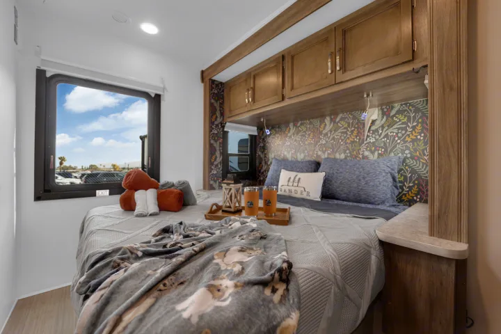 Experience comfort in this RV rental bedroom. Features a cozy bed, patterned wall, storage cabinets, and a window overlooking a parking area.. Jayco Redhawk 2021