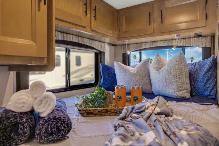 Cozy RV rental bedroom with patterned pillows, soft blankets, plant, and 'Let's Go Camping' tumblers. Palm trees visible through the window.. Coachmen Leprechaun 2021