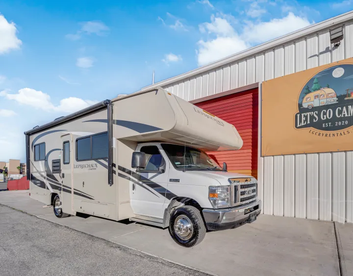 A cream-colored RV rental is parked by a building with a 'Let's Go Camping' sign. This motorhome is ready for your next adventure.. Coachmen Leprechaun 2021