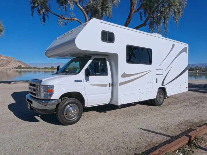 This white rv rental is parked lakeside with mountains and a tree overhead under a clear blue sky.. Winnebago Minnie Winnie 2020
