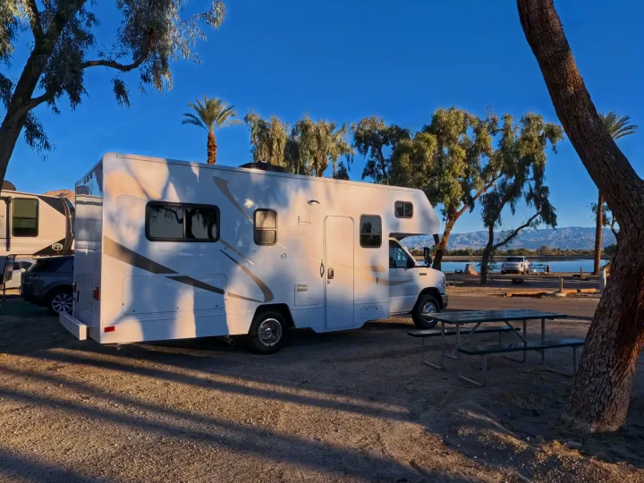 White RV rental at a sunny lakeside campsite with trees, distant mountains, and a picnic table by the water.. Winnebago Minnie Winnie 2020