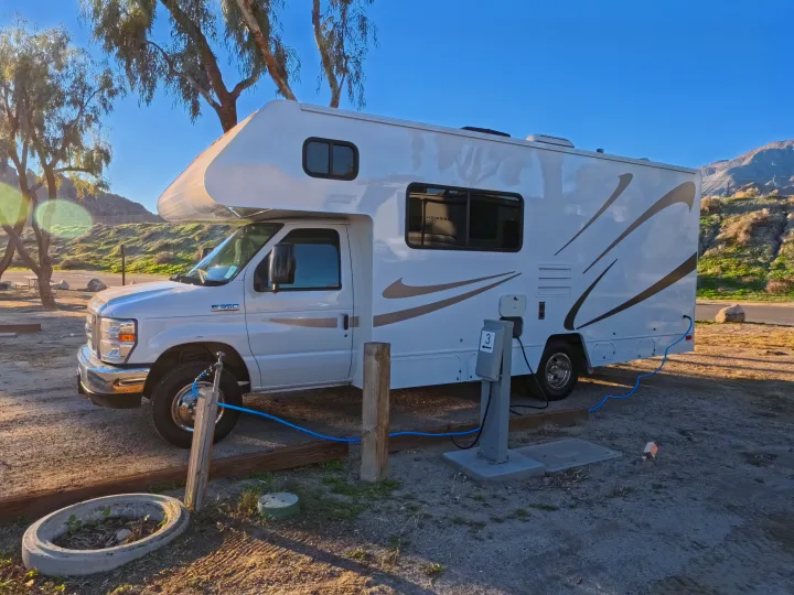 A white Ford E-350 rv rental is parked at a sunny campsite, connected to power and water, with mountains behind.. Winnebago Minnie Winnie 2020