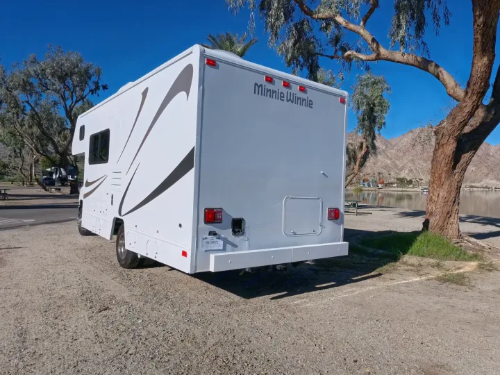A white Minnie Winnie RV rental is parked by a scenic lake with mountains under a clear blue sky, ready for an outdoor adventure.. Winnebago Minnie Winnie 2020