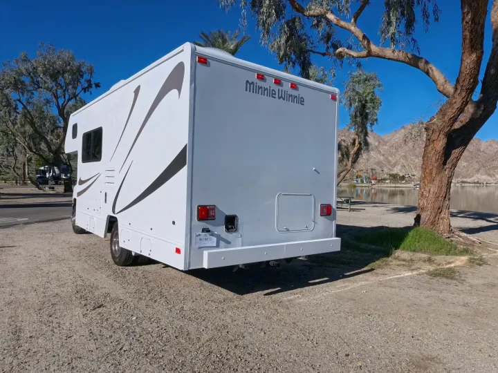 RV rental: Rear view of a white Minnie Winnie motorhome parked lakeside, surrounded by trees and mountains.. Winnebago Minnie Winnie 2020