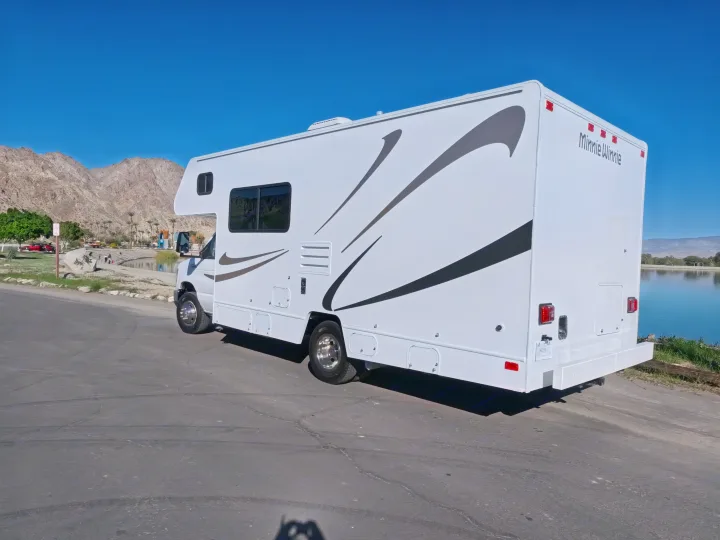 White Minnie Winnie RV rental with brown graphics parked by a lake, mountains, and clear blue sky.. Winnebago Minnie Winnie 2020