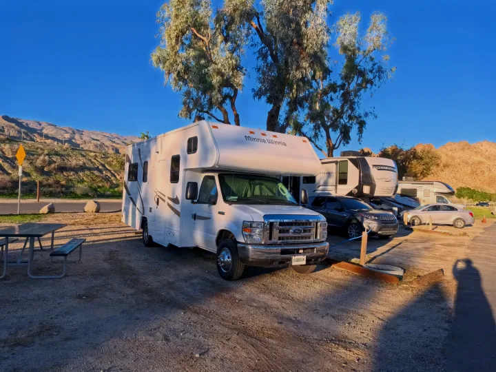 A white Minnie Winnie RV rental, marked Cruise America, parked at a sunny mountain campsite with a picnic table and scenic backdrop.. Winnebago Minnie Winnie 2020