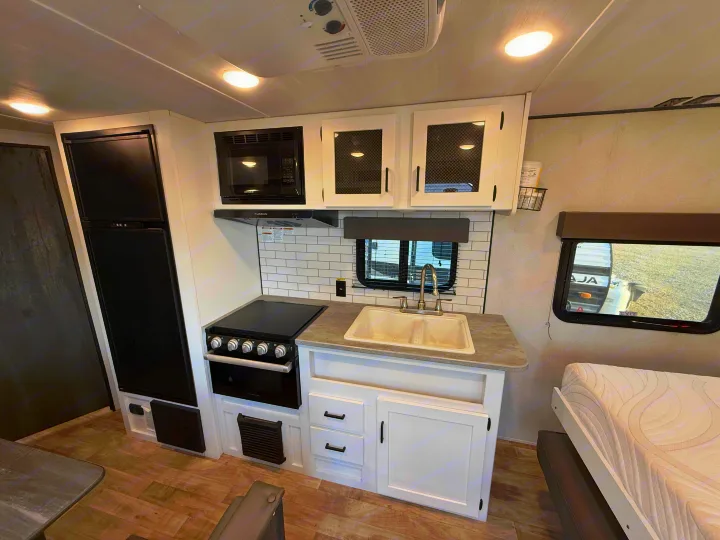 View of an rv rental kitchen featuring a black refrigerator, microwave, stove/oven, double sink, and white cabinets with a tiled backsplash.. Keystone Hideout 2022