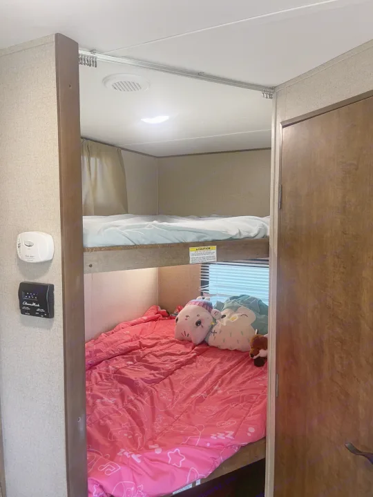 RV rental's bunk beds: a lower bed with pink bedding & toys, an upper bed. Wall has a smoke detector & thermostat.. Crossroads RV Zinger 2017