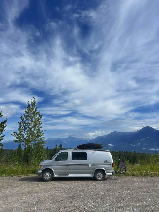 Silver RV rental van with bikes on rack, parked on a gravel road with scenic mountains under a cloudy sky.. Pleasure-Way Pleasure-Way Class B 2002