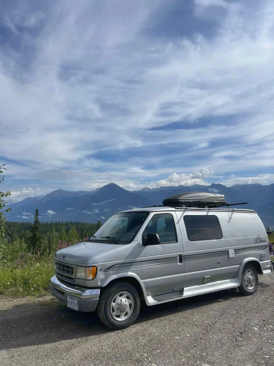 This scenic RV rental, a silver campervan with a roof box, is parked on a gravel path, showcasing breathtaking mountain views under a cloudy sky.. Pleasure-Way Pleasure-Way Class B 2002