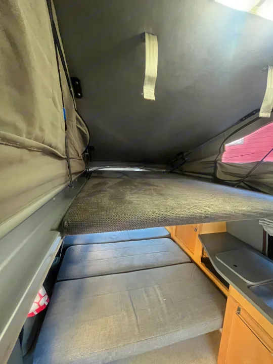 Inside this rv rental, an elevated pop-top sleeping bunk sits above lower cushions and a small kitchen counter.. Pleasure-Way Pleasure-Way Class B 2002