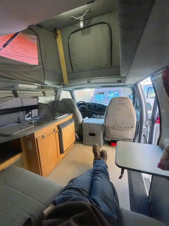 A person relaxing with feet up in a cozy rv rental camper van, featuring a pop-top roof and a compact kitchen. Travel comfort.. Pleasure-Way Pleasure-Way Class B 2002