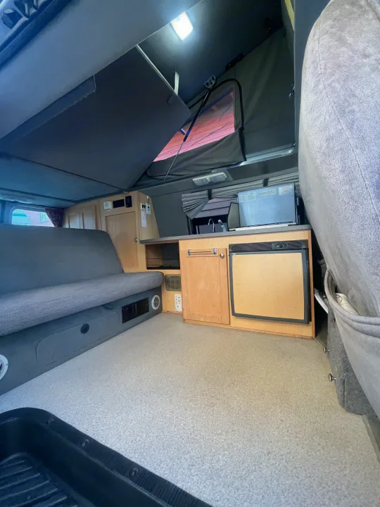 Spacious RV rental interior with a pop-top, convertible sofa bed, and compact kitchenette featuring a fridge, sink, and cabinets.. Pleasure-Way Pleasure-Way Class B 2002