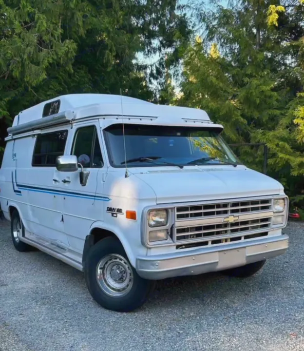 White vintage Chevy camper van RV rental with high top & awning, parked on gravel amidst green trees. Ready for travel adventures.. Chevrolet G20 1987