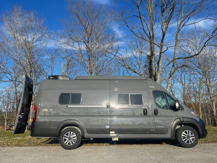 A gray Winnebago Solis RV rental is parked on pavement with its rear door open, set against bare trees and a clear blue sky.. Winnebago Solis 2025