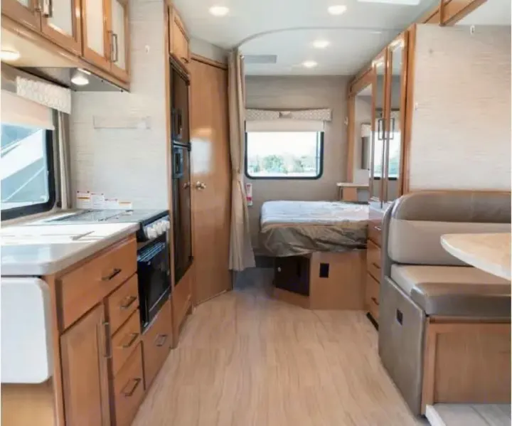 Bright rv rental interior: kitchen on left, bed by window, and dining booth on right. Features light wood tones.. Thor Quantum 2021