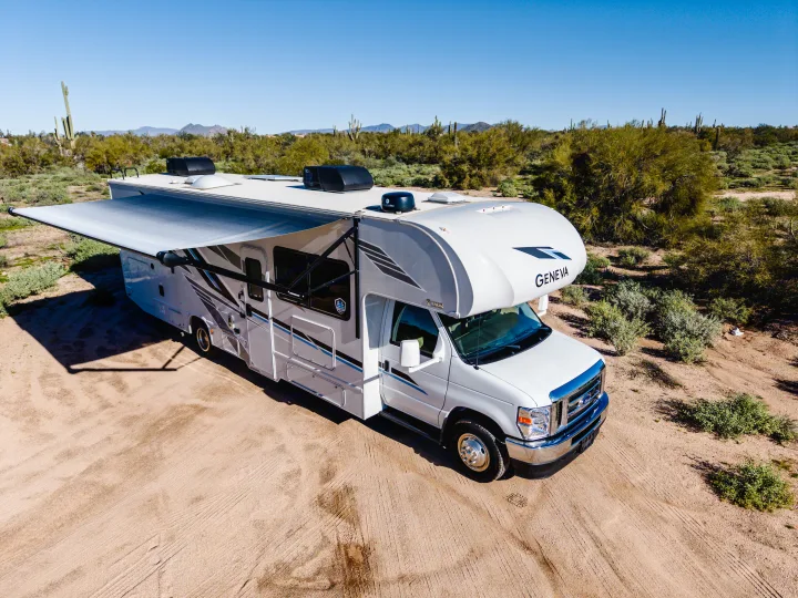 Aerial view of a white Geneva RV rental with its awning extended, parked on a dirt lot in a sunny desert with cacti.. Thor Geneva 2025