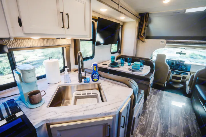 Discover this modern RV rental interior, showcasing a bright kitchen with marble counters, cozy dining booth, and cockpit view.. Thor Geneva 2025