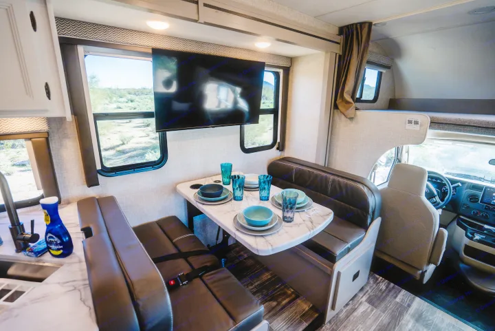 Inside this RV rental: a cozy dinette, kitchen sink, TV, and cab-over bunk, with desert landscape views.. Thor Geneva 2025