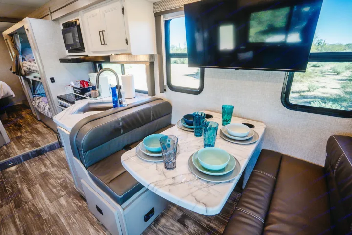 Modern RV rental interior: dinette with place settings, TV, fully-equipped kitchen with microwave/sink, comfy bunk beds, and scenic desert views.. Thor Geneva 2025