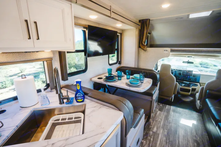 Inside a comfortable rv rental: a well-lit kitchen, dining space with place settings, and a view of the driver's area.. Thor Geneva 2025